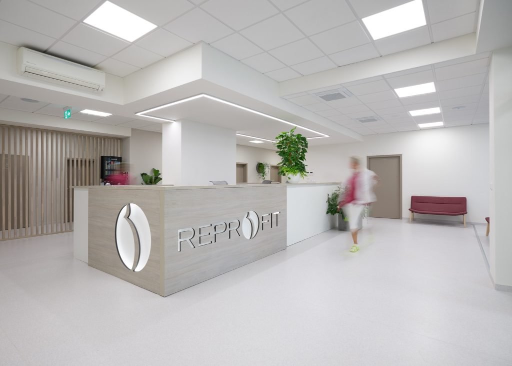 Modern reception desk with REPROFIT branding, white-and-wood finish, plants, and a blurred visitor in a bright medical lobby.